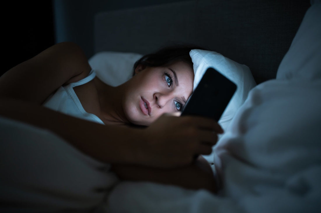 A woman laying in bed lit only by the light of the cell phone in her hand.