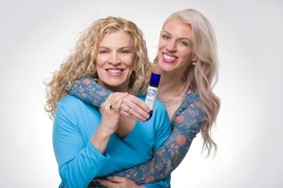 Two woman with their arms around one another, holding a bottle of a Vitali Skin Care product.